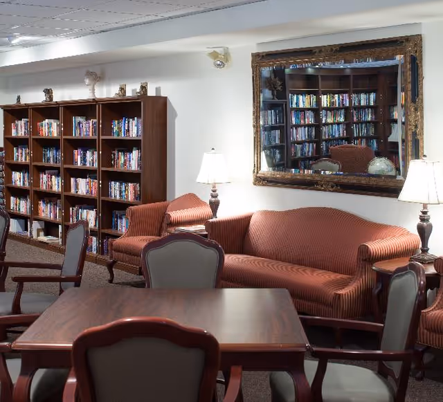 A cozy library or reading room with wooden bookshelves filled with books, a large mirror on the wall reflecting the shelves, two table lamps on side tables, a striped upholstered sofa and armchairs, and a wooden table with chairs in the foreground.