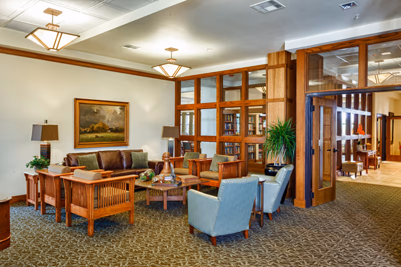 A cozy senior living facility common area with wooden framed furniture including a brown leather sofa, wooden armchairs with cushions, and two light blue upholstered chairs. The room features a patterned carpet, two table lamps on side tables, a framed landscape painting on the wall, and large wooden-framed glass windows and doors leading to another room with bookshelves.