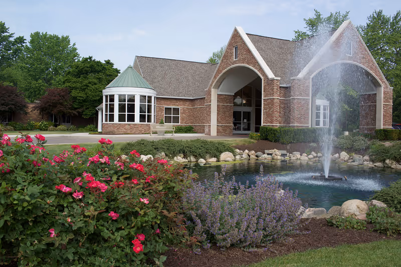 Brick senior living facility entrance with a fountain and landscaped flower beds in front.