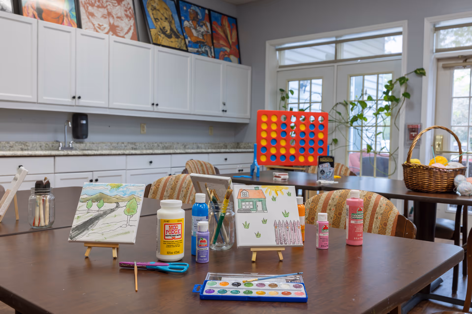 A bright activity room with tables and chairs featuring art supplies including paint bottles, brushes, scissors, and watercolor palettes. Two small canvases with paintings are displayed on the table. In the background, there is a large Connect Four game, a basket with yarn, and large windows letting in natural light. White cabinets with colorful artwork above them line the wall.
