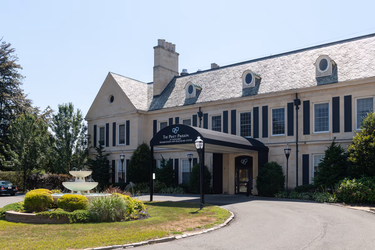 Exterior view of a large, elegant two-story building with a stone facade and black shutters. The entrance features a black canopy with the text 'The Pratt Pavilion at Glengariff Rehabilitation and Healthcare Center.' There is a circular driveway with a landscaped island containing a tiered fountain and various shrubs and plants. Trees and greenery surround the building under a clear blue sky.