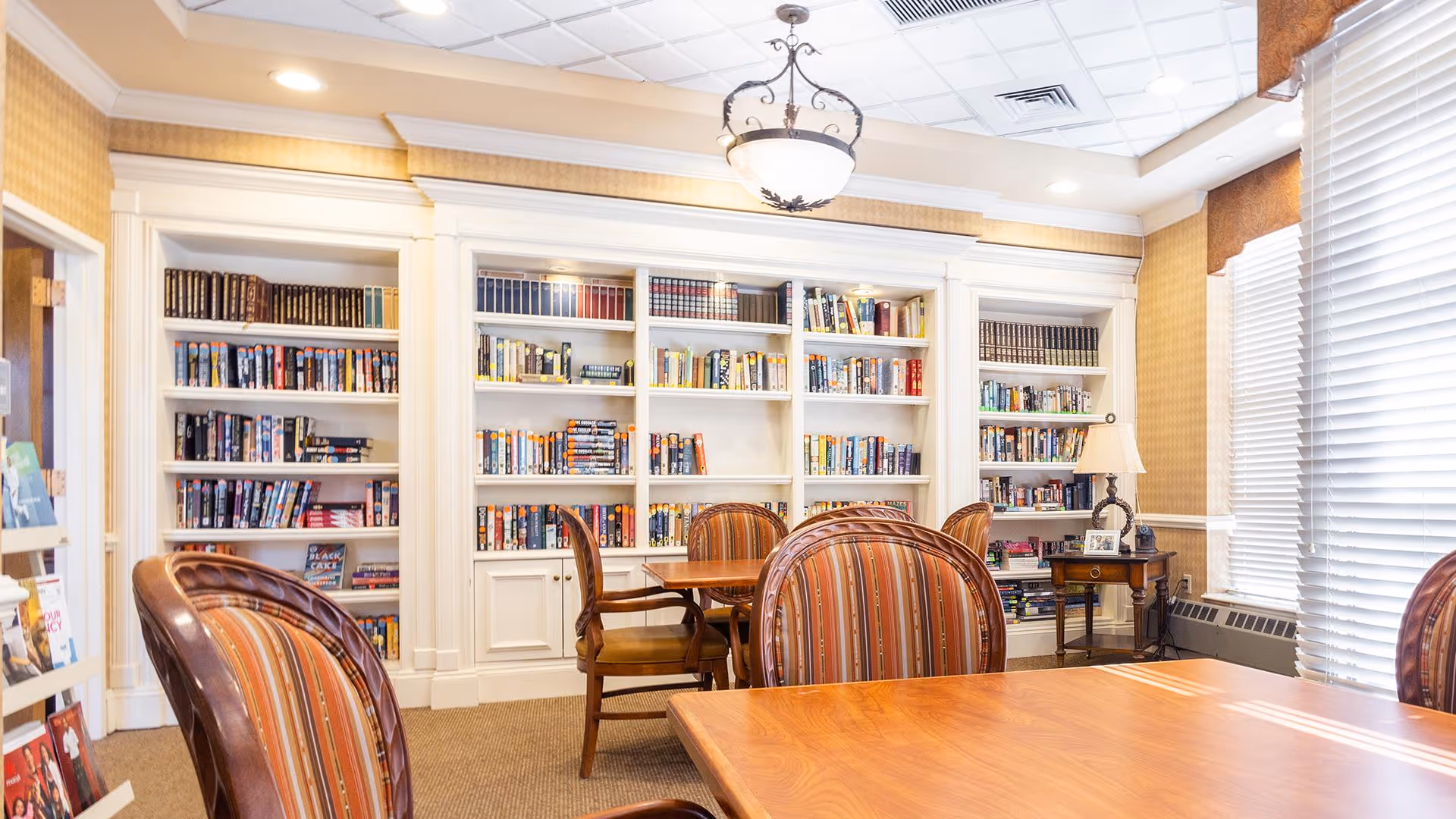 A well-lit library room with built-in white bookshelves filled with books. There are wooden tables and striped upholstered chairs arranged for reading or small group activities. A window with blinds allows natural light to enter the room, and a decorative ceiling light fixture hangs above.