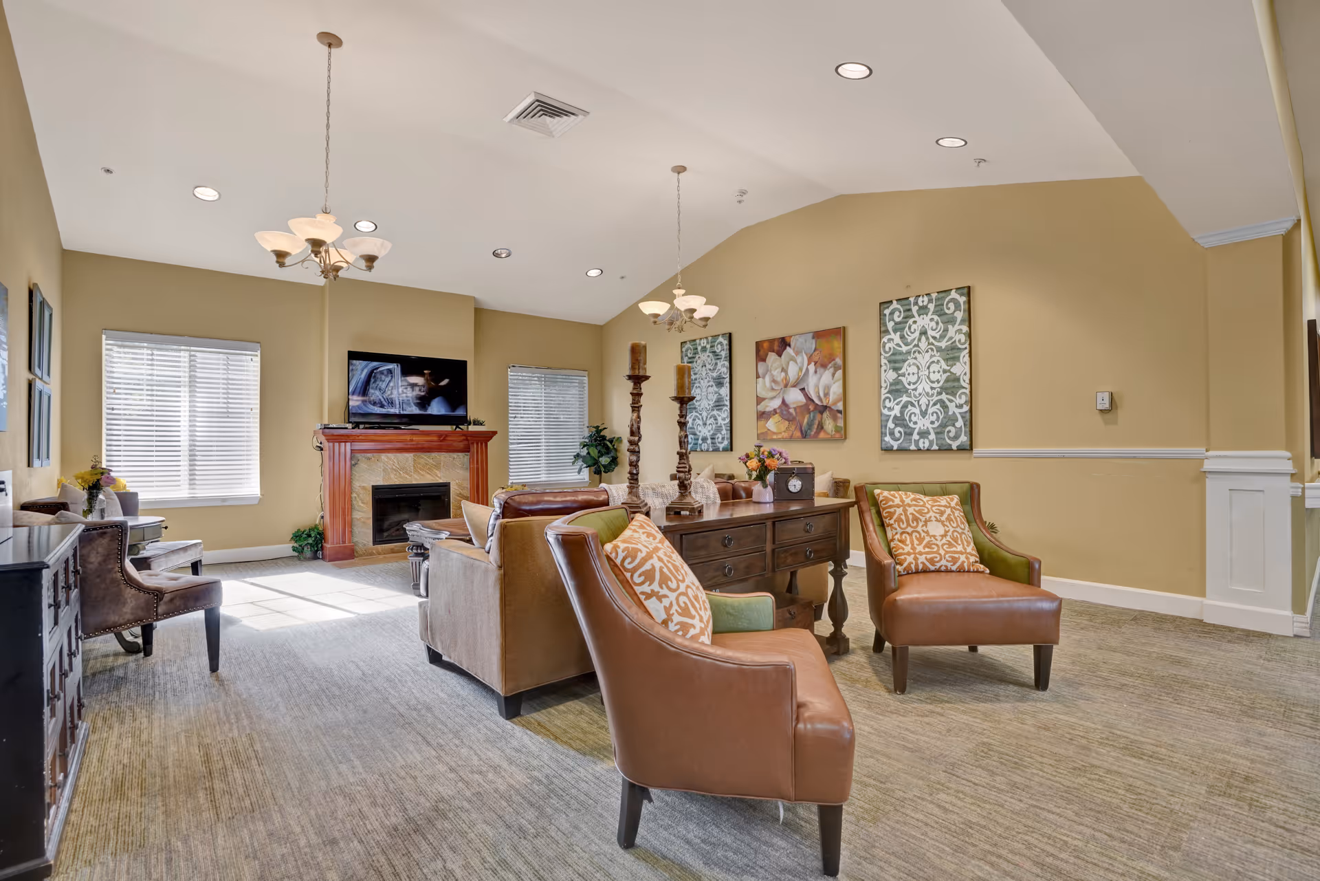 A cozy living room area with beige walls and carpeted floor. The room features a fireplace with a TV mounted above it, two windows with blinds, and comfortable seating including two brown leather chairs with patterned cushions and a sofa. There is a wooden console table with decorative items and wall art above it. Ceiling lights and a chandelier provide illumination.