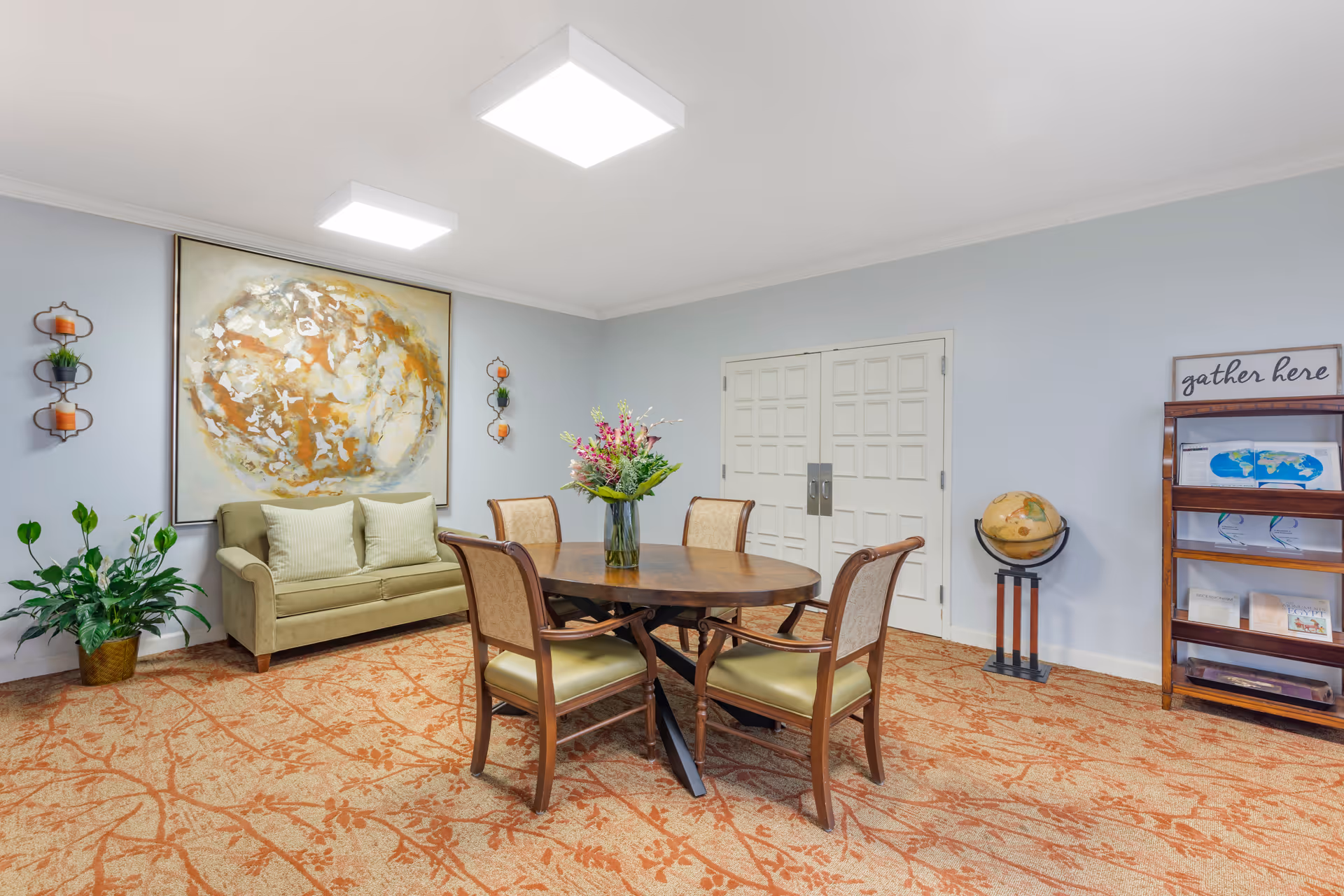 A furnished community room with a round wooden table and chairs, a green loveseat, large abstract wall art, potted plants, and a bookshelf.