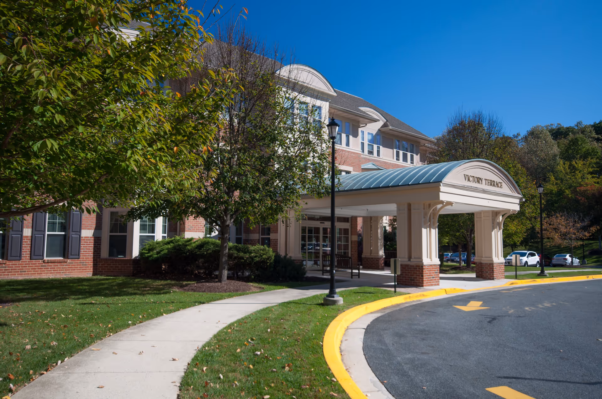 Exterior view of Victory Terrace senior living facility showing the entrance with a covered driveway, surrounded by trees and a curved sidewalk on a sunny day.