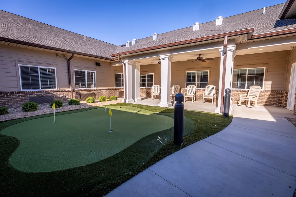 Outdoor courtyard area with a small putting green featuring three yellow flags, surrounded by a concrete walkway. The building has beige siding with brick accents and several windows. There is a covered patio with white columns and several wooden chairs arranged along the wall. The sky is clear and blue.