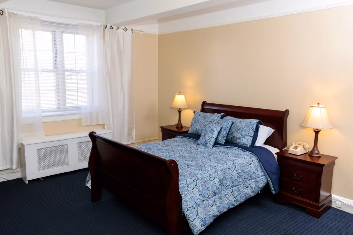 Sunlit bedroom with a dark wood sleigh bed dressed in blue patterned bedding, flanked by matching nightstands and lamps.