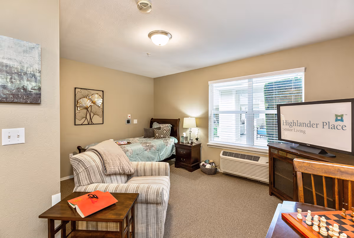 A cozy senior living room featuring a single bed with floral bedding, a striped armchair with a beige throw, a wooden side table with a red book and glasses, a nightstand with a lamp and decor, a window with blinds, an air conditioning unit below the window, a TV on a wooden stand displaying the Highlander Place Senior Living logo, and a chessboard on a wooden table in the foreground.