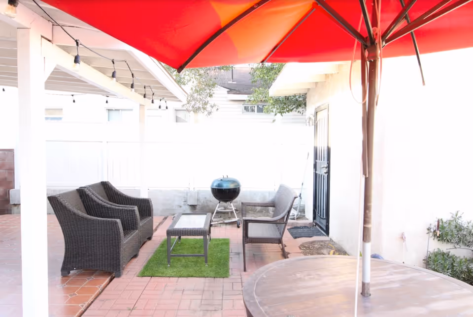Outdoor patio area with a red umbrella, brown wicker chairs, a glass-top coffee table on a small green rug, a round table in the foreground, a black charcoal grill, and string lights hanging from a white pergola.