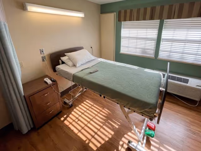Sunlit patient room with an adjustable hospital bed covered by a green blanket, a bedside cabinet, windows with blinds, and an air conditioner.