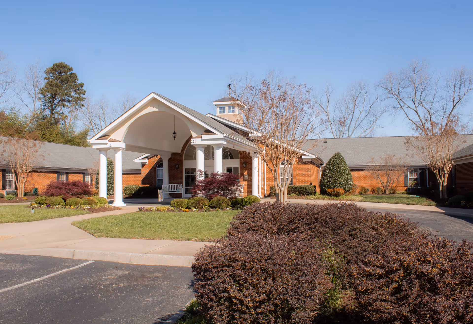 Front exterior view of a single-story brick building with a covered entrance supported by white columns. The building is surrounded by landscaped bushes, trees, and a paved driveway under a clear blue sky.