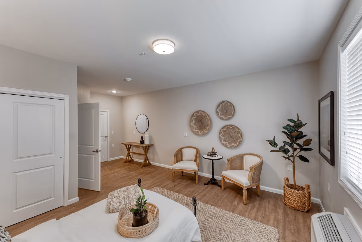A bright and cozy bedroom in Cedarhurst Senior Living of Owensboro featuring a bed with a beige throw blanket and a small plant on a tray. The room has wooden flooring, two wooden chairs with white cushions, a small round table with books and a decorative bird, a woven basket with a tall plant, three decorative woven wall hangings, a wooden console table with a round mirror above it, and a window with white blinds letting in natural light.