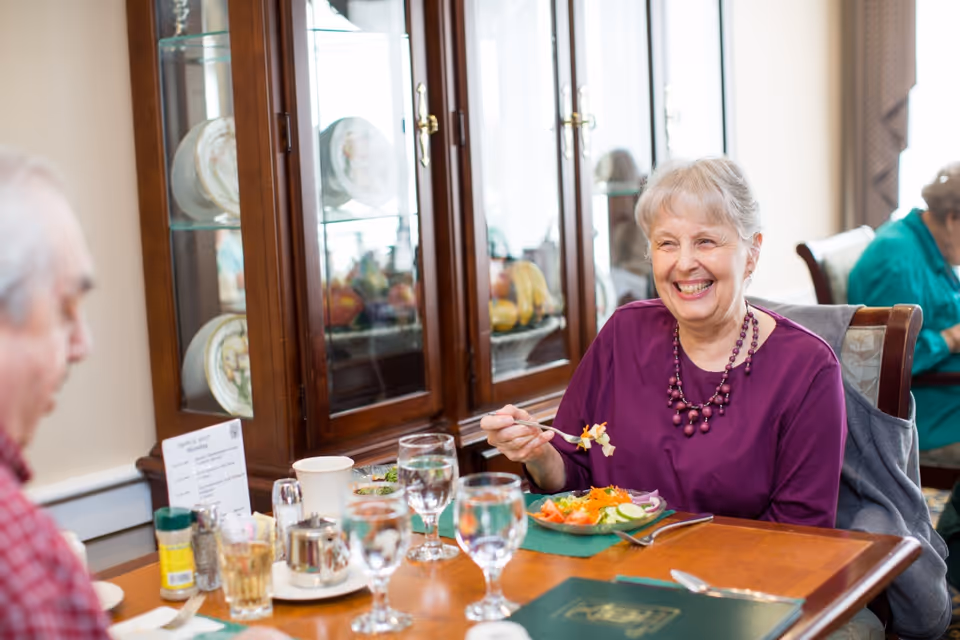 An elderly woman wearing a purple top and necklace is smiling and holding a fork with food while sitting at a dining table with plates of salad, glasses of water, and condiments. Another elderly person is partially visible sitting across from her. In the background, there is a wooden cabinet with glass doors displaying plates and fruit.