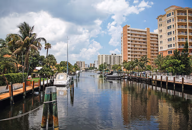 A canal with docked boats lined by palm trees and multi-story residential buildings under a partly cloudy sky.