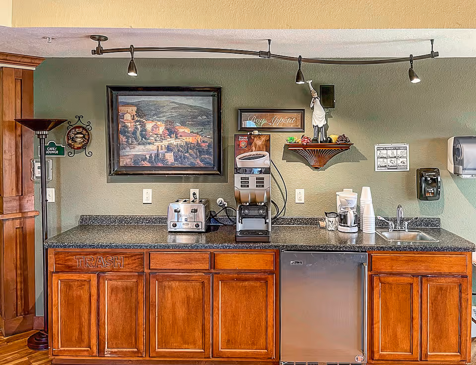 A kitchenette area with wooden cabinets and a black countertop. On the countertop, there is a toaster, a coffee machine, a small coffee maker, disposable cups, and a sink. Above the counter, there is a framed landscape painting, a 'Bon Appetit' sign, a decorative chef figurine on a wall shelf, and a handwashing reminder poster. The wall is painted green, and there is a floor lamp and a small sign that reads 'Cafe Lounge' on the left side.