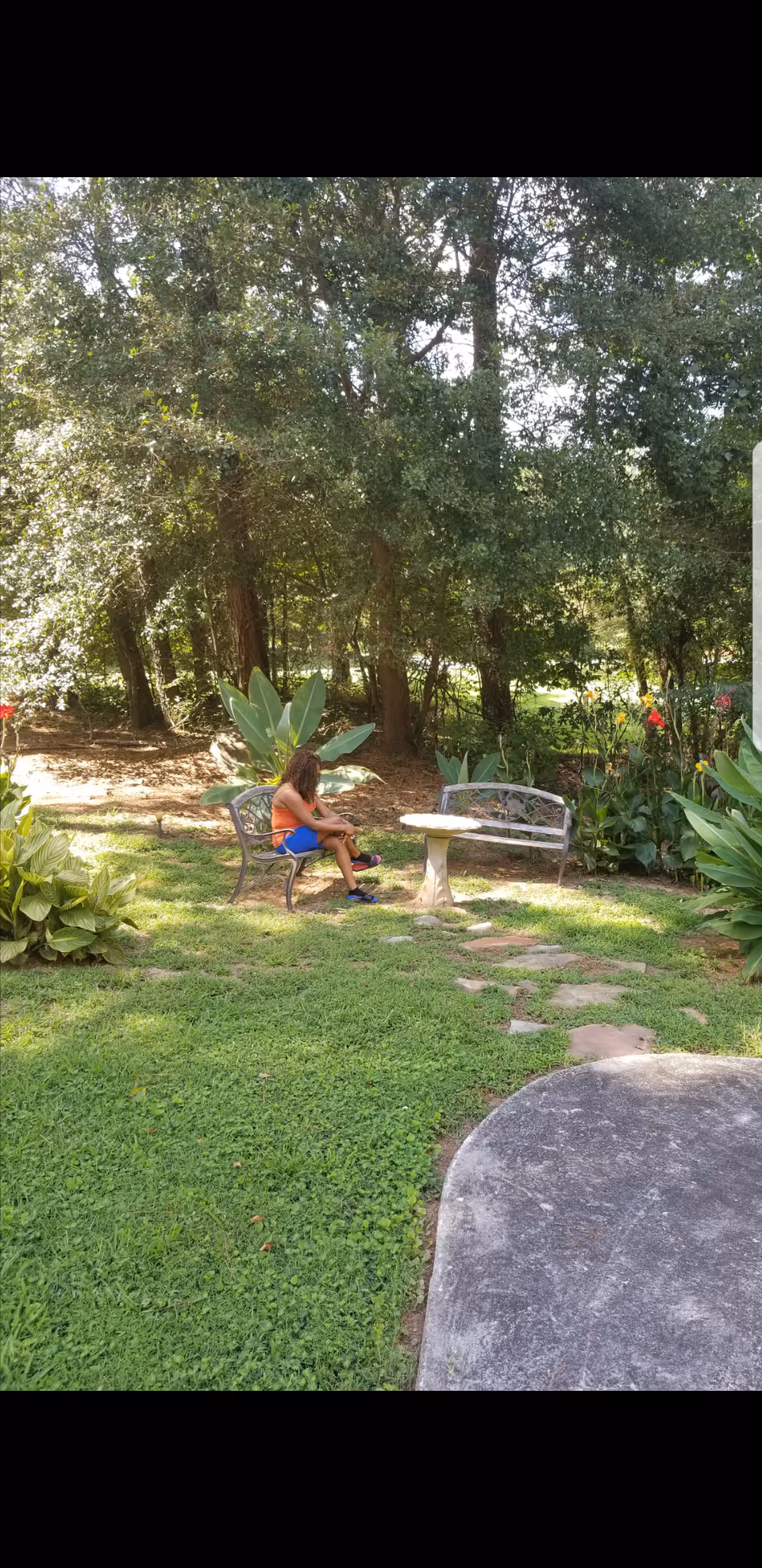 A person sits on a bench beside a small round table in a shaded garden area with trees and plants.