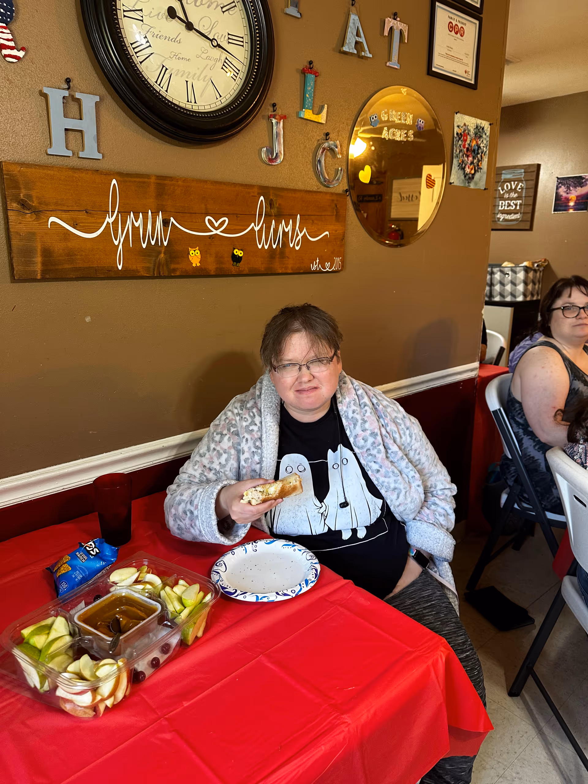 A woman wearing glasses and a patterned robe sits at a table with a red tablecloth, holding a sandwich. On the table is a tray with apple slices, celery, and a container of caramel dip, along with a bag of chips and a paper plate. Behind her, the wall is decorated with a large clock, colorful letters, a wooden sign that says 'Green Acres,' a round mirror, and various framed pictures and signs. Another woman is seated in the background.