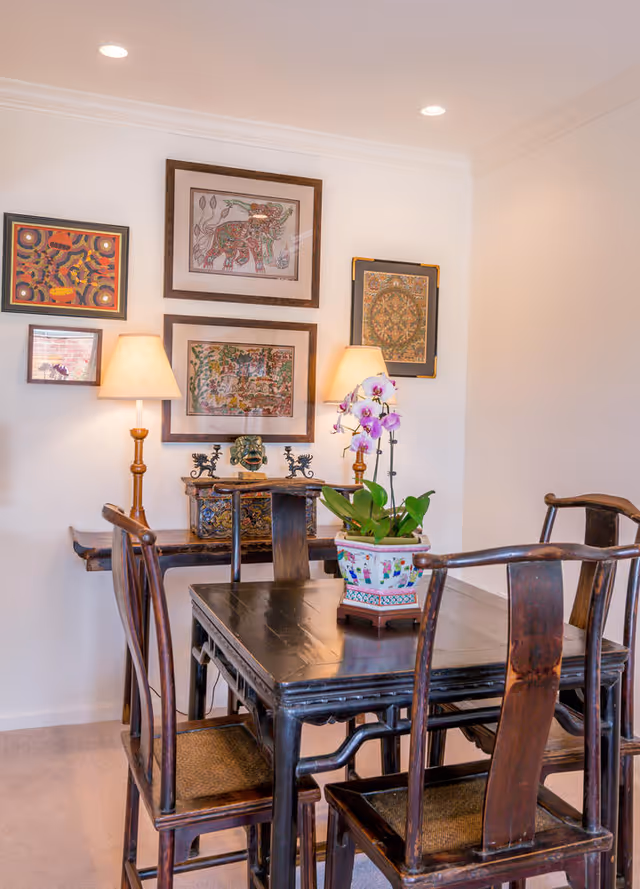 Small dining area with a dark wood table and chairs, a potted orchid centerpiece, two lamps, and framed artwork on the wall.