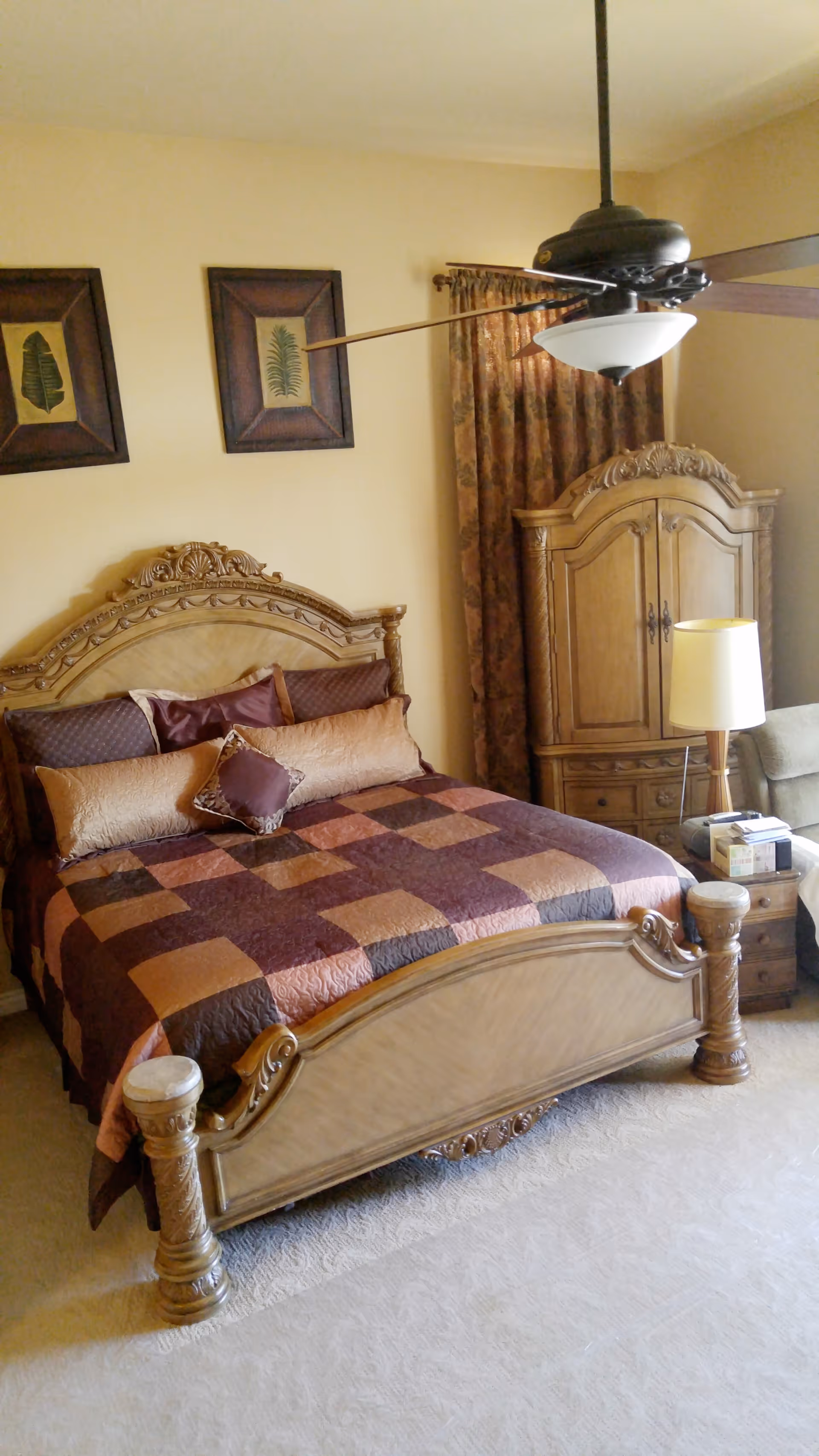 A bedroom with a large wooden bed featuring an ornate headboard and footboard. The bed is covered with a patchwork quilt in shades of brown and orange, and several pillows. Behind the bed are two framed botanical prints on a beige wall. To the right of the bed is a wooden armoire and a nightstand with a lamp and some items on it. A ceiling fan with a light fixture hangs above the bed. The room has beige carpet and curtains with a floral pattern.