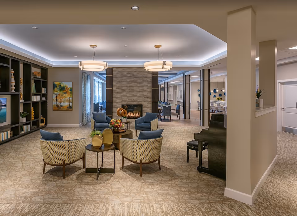 A spacious and elegantly decorated common area in a senior living facility featuring a seating arrangement with four armchairs around a small table with decorative items, a modern fireplace with a stone surround, built-in shelving with books and decor, a piano on the right side, and a dining area visible in the background.
