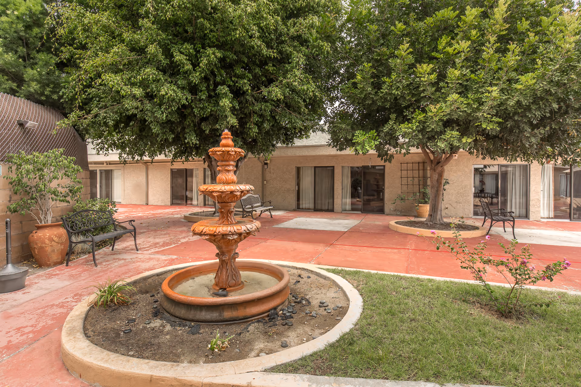 Outdoor courtyard area with a three-tiered terracotta fountain in the center, surrounded by a circular planter with some plants. There are benches placed around the courtyard, large trees providing shade, and sliding glass doors leading into the building.