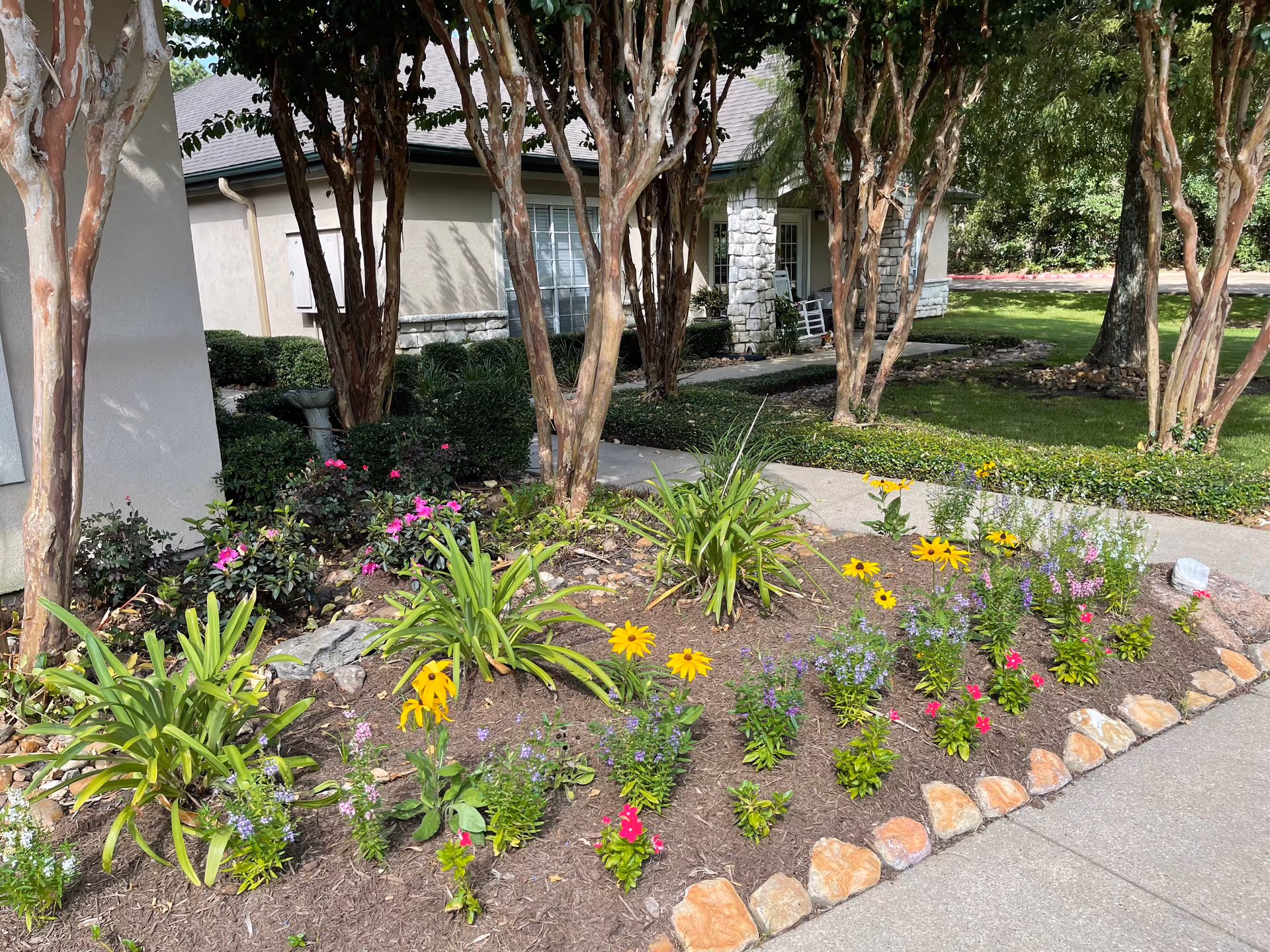 A landscaped garden area with colorful flowers and green plants bordered by rocks, situated next to a concrete walkway. In the background, there are several trees and a building with stone and stucco exterior walls, windows, and a porch with a rocking chair.
