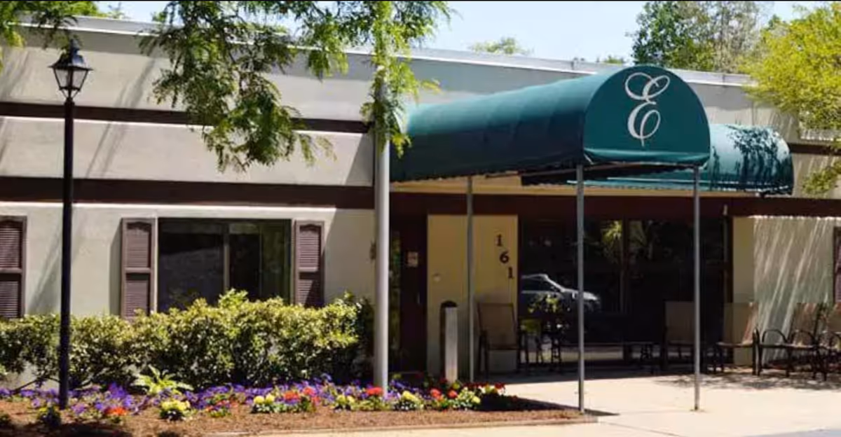 Exterior view of a building entrance with a green awning displaying a stylized letter E. The building has beige walls with brown trim, windows with brown shutters, and a landscaped area with bushes and colorful flowers in front. There are chairs placed near the entrance under the awning.