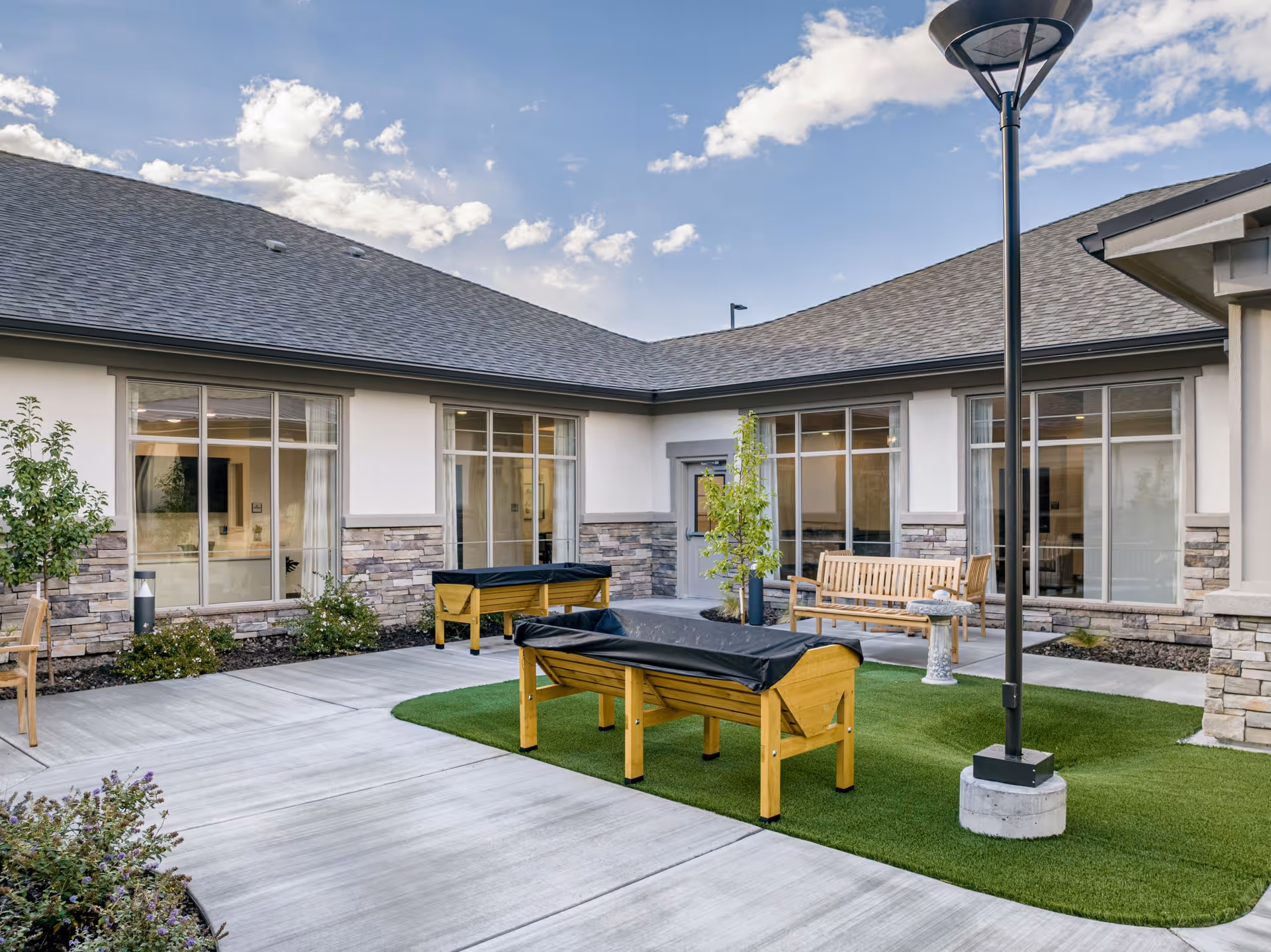 Outdoor courtyard area of Kiley Ranch Senior Living with a concrete walkway, artificial grass patches, wooden benches, covered raised garden beds, small trees, and a tall lamp post under a partly cloudy sky.