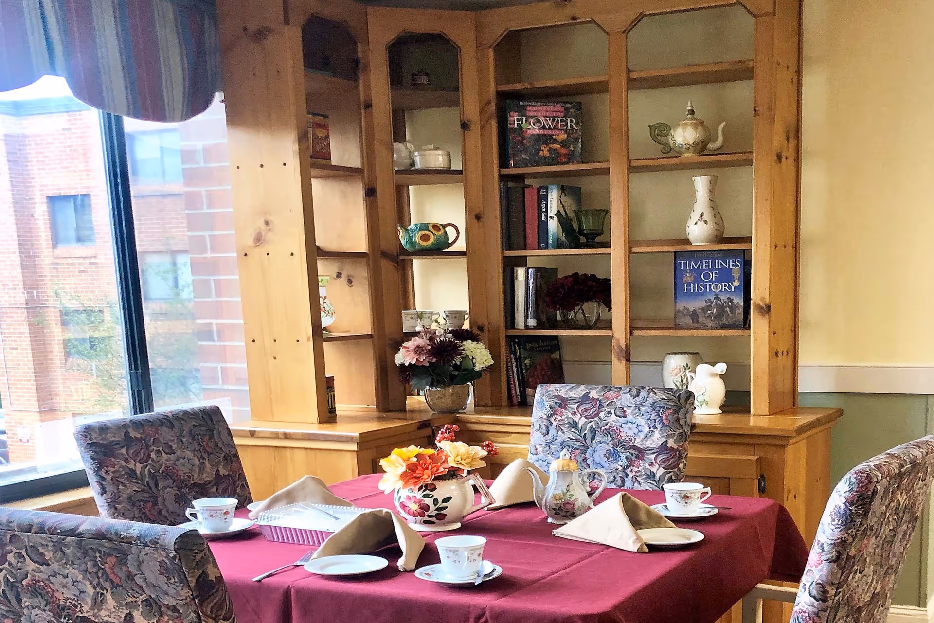 A dining area with a table covered in a red tablecloth set for tea with floral-patterned chairs around it. The table has a floral centerpiece, teacups, saucers, and folded napkins. Behind the table is a wooden bookshelf with decorative items and books. A window with a striped valance lets in natural light.