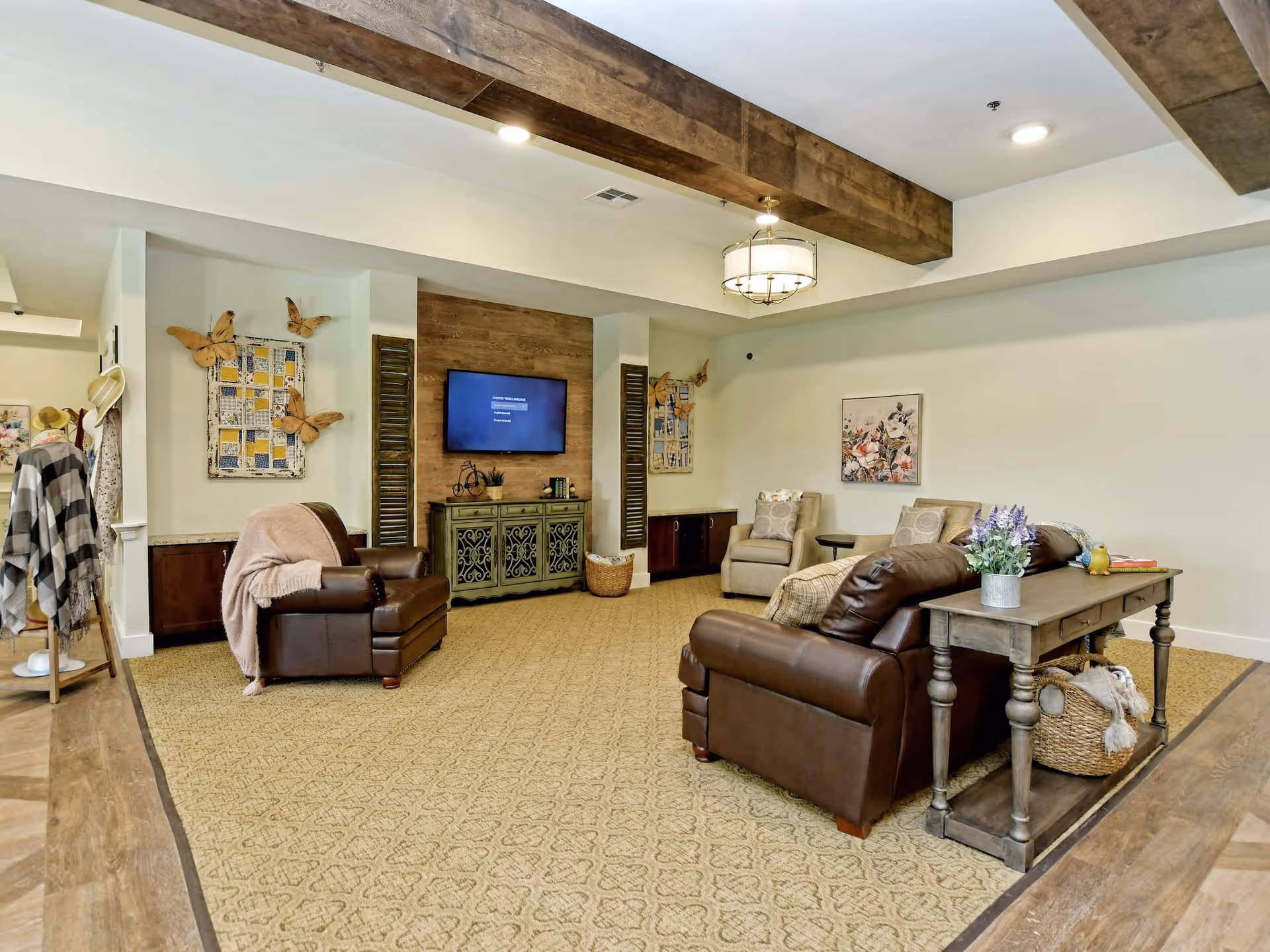 A cozy living room area with beige carpet and wooden ceiling beams. The room features two brown leather armchairs, a brown leather sofa, and two beige upholstered chairs. A wooden console table with a basket underneath and a vase with flowers is behind the sofa. A flat-screen TV is mounted on a wooden accent wall above a decorative cabinet. The walls are decorated with butterfly wall art and a floral painting. The room has soft, warm lighting and a comfortable, inviting atmosphere.