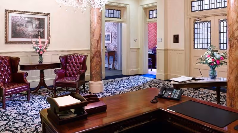 An elegant interior room with a wooden reception desk in the foreground, featuring a telephone and office supplies. Behind the desk, there are two burgundy leather armchairs with a small round table holding a vase of flowers. The room has patterned carpet, cream-colored walls with wainscoting, two large decorative columns, and stained glass windows above the doorways. There are open doorways leading to other rooms with floral wallpaper and additional furniture.