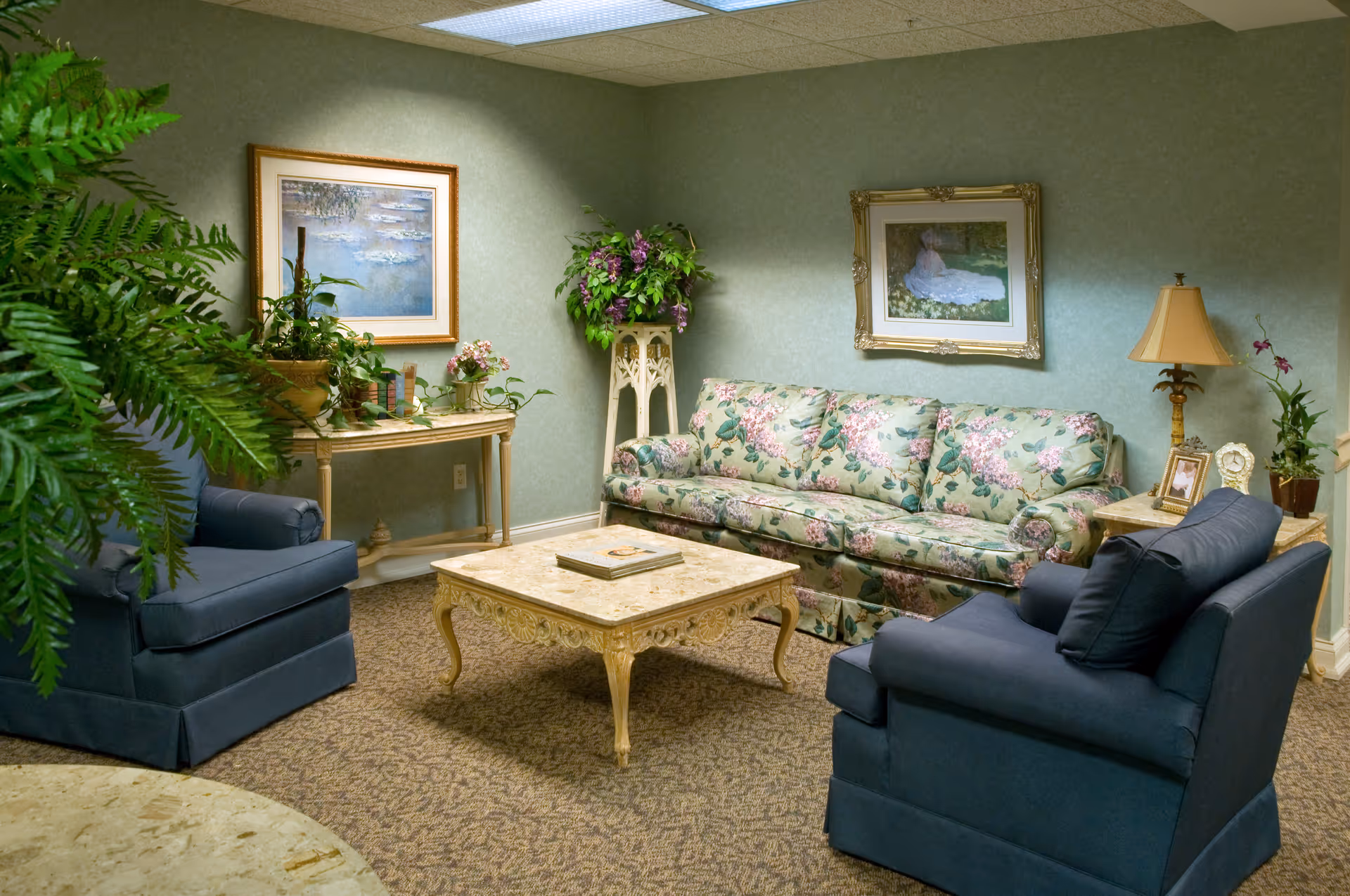 A cozy sitting room with a floral sofa, two blue armchairs, a carved coffee table, and decorative plants and artwork on the walls.