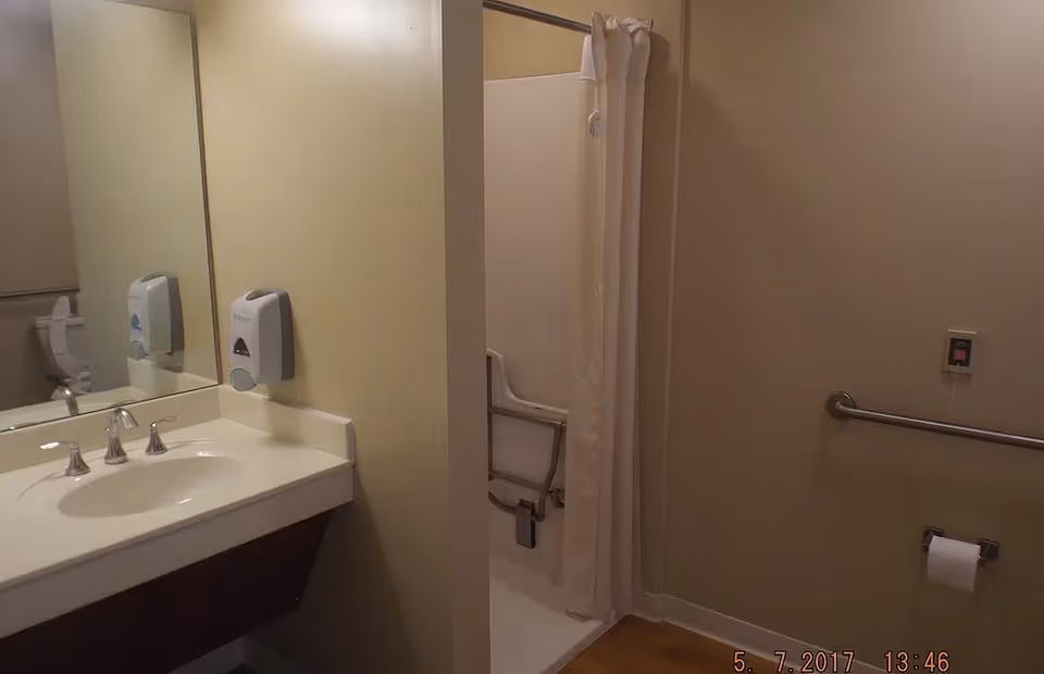 A bathroom with a sink and faucet on the left, a large mirror above the sink, a soap dispenser mounted on the wall, a shower area with a white curtain and grab bars, a toilet paper holder with a roll of toilet paper, and a grab bar on the right wall. The walls are painted beige and the floor appears to be wood or wood-like.