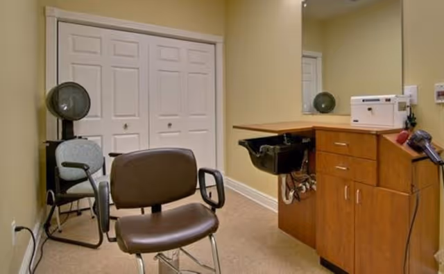 Interior of a small hair salon or beauty station with a brown salon chair in the foreground, a hair dryer chair in the back left corner, a wooden cabinet with drawers and a black sink, a hair dryer attached to the cabinet, and a large mirror on the wall.