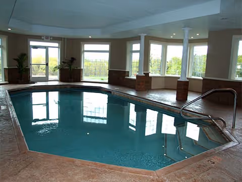 Indoor swimming pool with clear blue water surrounded by a tiled deck, large windows letting in natural light, and potted plants near the exit door.