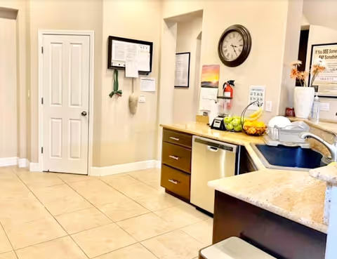 Interior view of a clean kitchen area in a senior care facility featuring beige tiled floors, a countertop with a sink, a dishwasher, a bowl of fruit, a wall clock, and various notices and decorations on the walls.