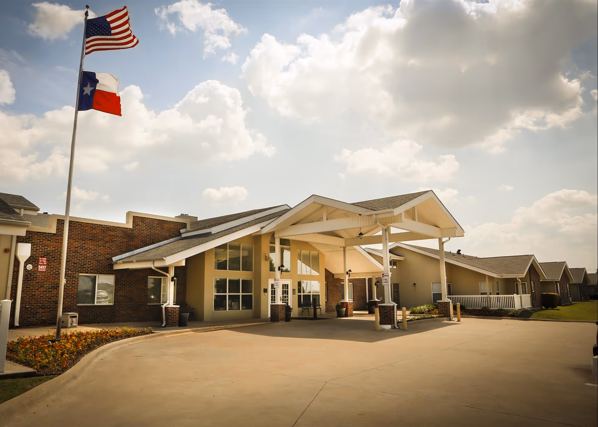 Exterior view of Bethesda Gardens Fort Worth senior living facility showing the main entrance with a covered driveway, American and Texas flags flying on a flagpole, and a partly cloudy sky.