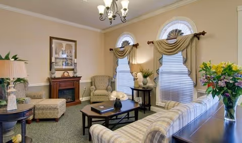 A cozy living room with a striped sofa, an armchair, a coffee table with decorative items, a small fireplace with framed artwork above it, two arched windows with curtains, and a chandelier hanging from the ceiling.