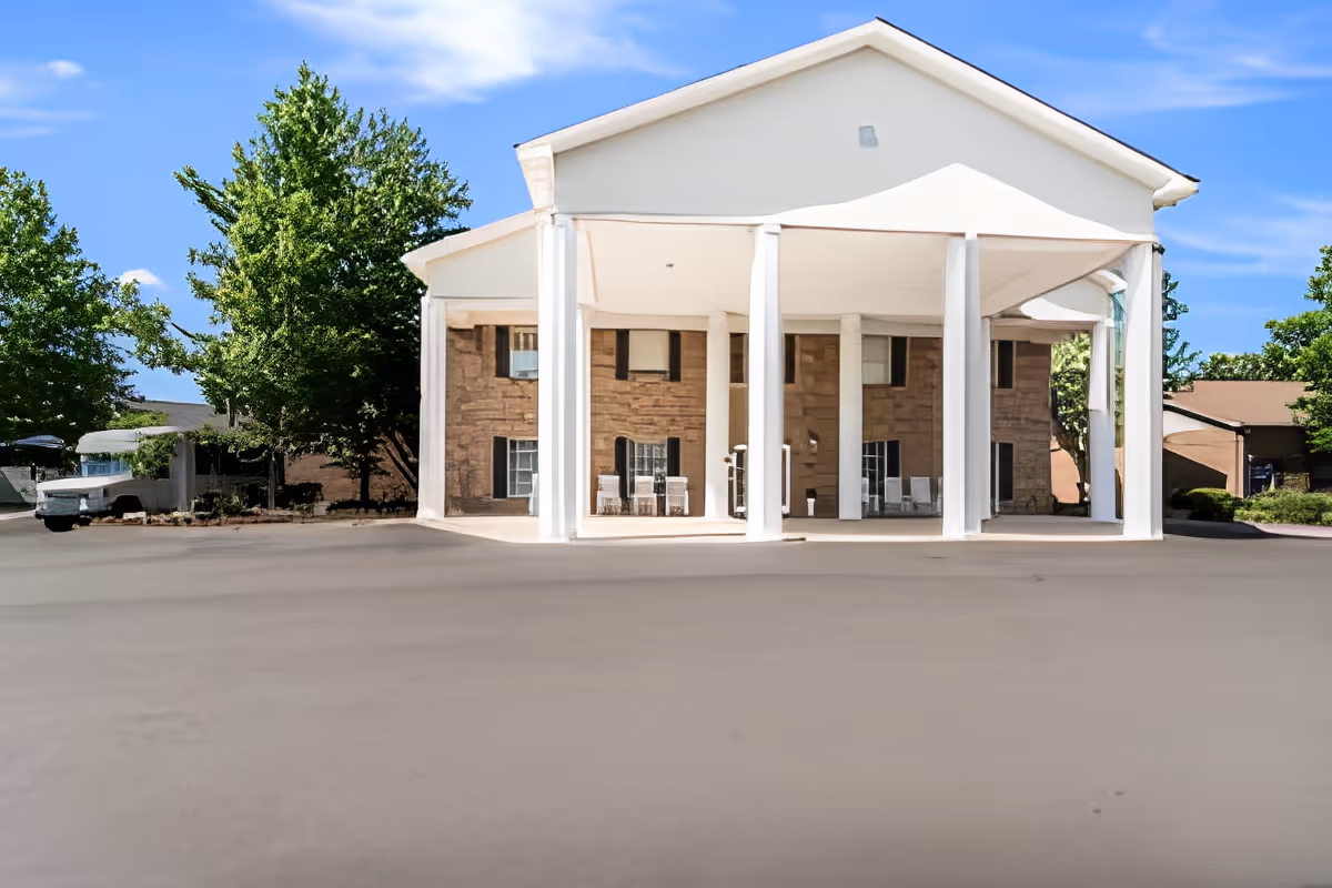 Front exterior view of a building with large white columns supporting a covered entrance area, surrounded by trees and clear blue sky.