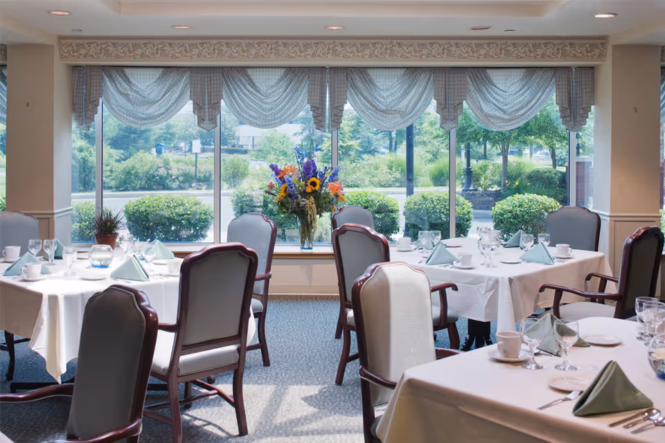 A bright dining room with several tables covered in white tablecloths, each set with folded green napkins, glassware, cups, and plates. The room has large windows with sheer curtains, allowing natural light to illuminate the space. A vase with colorful flowers is placed on the windowsill, and greenery is visible outside.