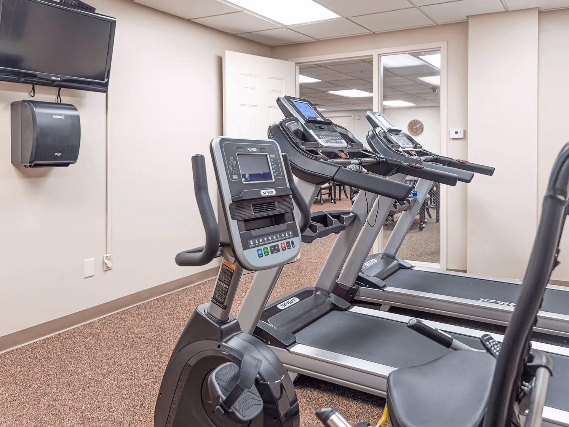 Indoor fitness room with exercise equipment including a stationary bike and two treadmills, a wall-mounted TV, a paper towel dispenser, and a large mirror reflecting part of the room.