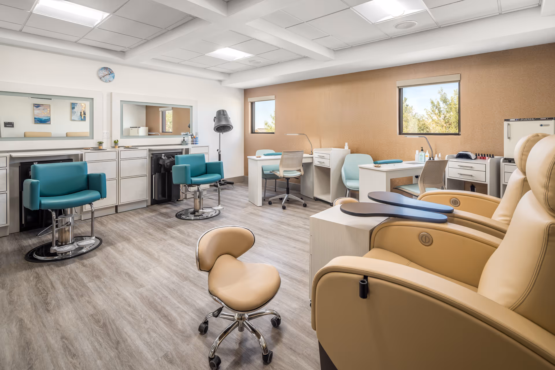 A bright and clean salon and manicure area in a senior living facility featuring two teal salon chairs in front of large mirrors, beige pedicure chairs with attached footrests, and manicure tables with chairs and nail care supplies. The room has wood-look flooring, white walls, and two windows letting in natural light.