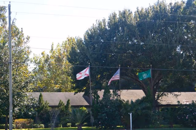 Single-story facility front with three flags (Florida, U.S., and a green flag) flying among trees and landscaping.