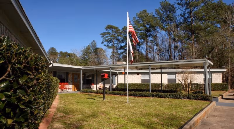 Single-story brick building with a covered walkway and an American flag on a flagpole in the front yard, surrounded by grass, bushes, and trees under a clear blue sky.