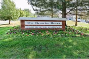 Brick entrance sign that reads 'The Beatrice Hover Assisted Living Residence' on a grassy lawn with trees and a road behind it.