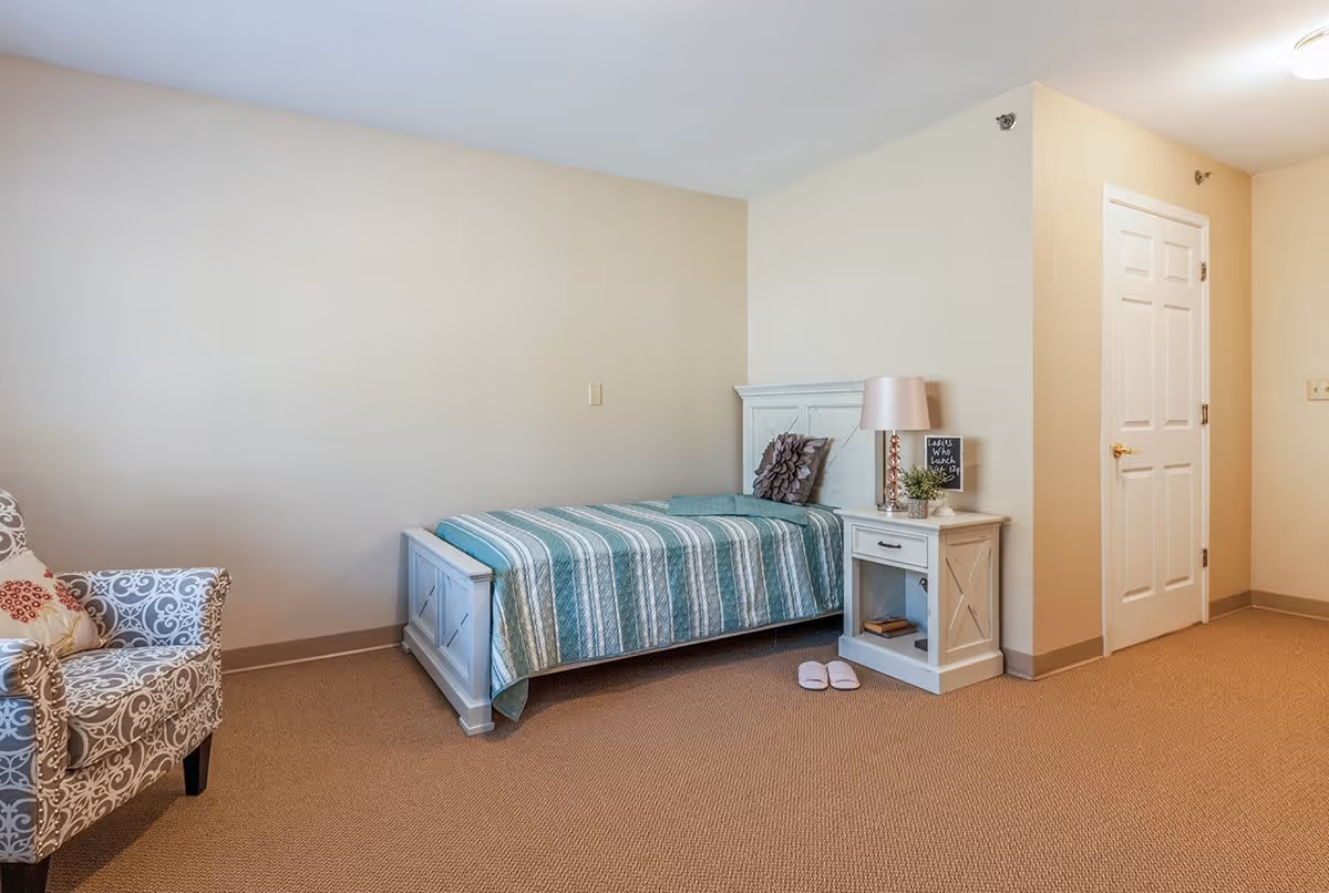 A simple bedroom with a single bed covered in a green and white striped quilt, a white nightstand with a lamp, small plant, and decorative items, a patterned armchair with a cushion, beige carpet, and a closed white door.
