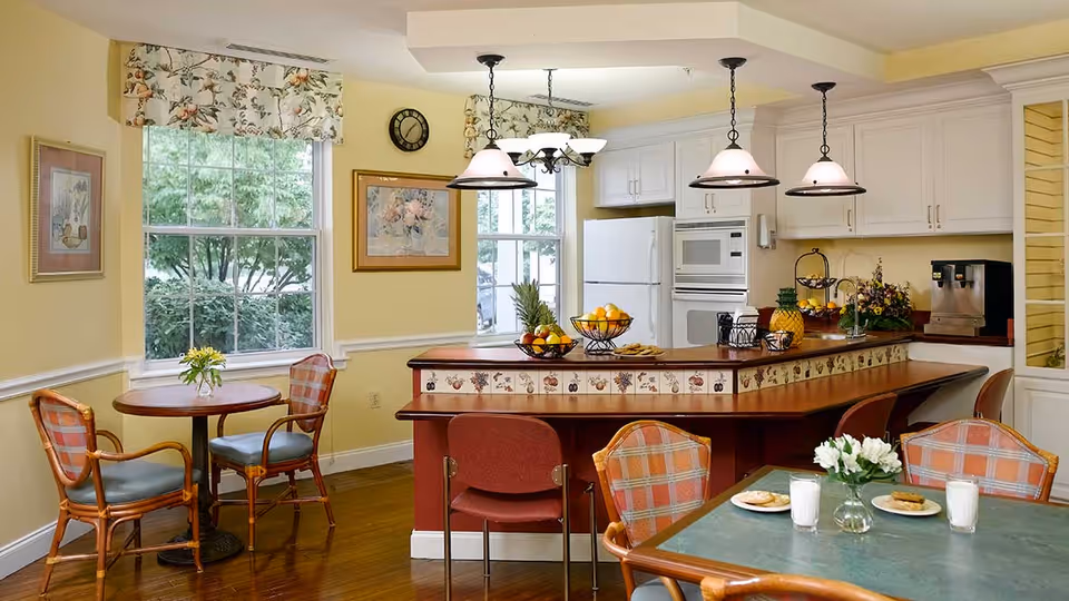 A bright and cozy kitchen and dining area with yellow walls and wooden flooring. The kitchen features white cabinets, a refrigerator, microwave, and a coffee machine. A curved counter with decorative tiles and three hanging pendant lights separates the kitchen from the dining area. There are two small round tables with chairs, one near the window and one in the foreground with plates of cookies and glasses of milk. Floral curtains adorn the windows, and framed artwork and a clock hang on the walls.