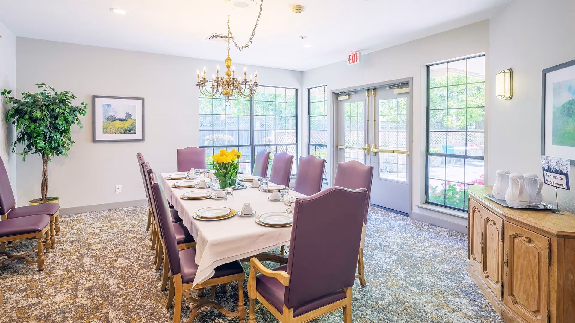 A bright dining room with a long table set for a meal, featuring purple cushioned chairs, a white tablecloth, plates, cups, and a centerpiece of yellow flowers. Large windows and glass doors let in natural light, and a chandelier hangs above the table. A wooden sideboard with pitchers and a sign is against one wall, and a potted plant and framed artwork decorate the room.