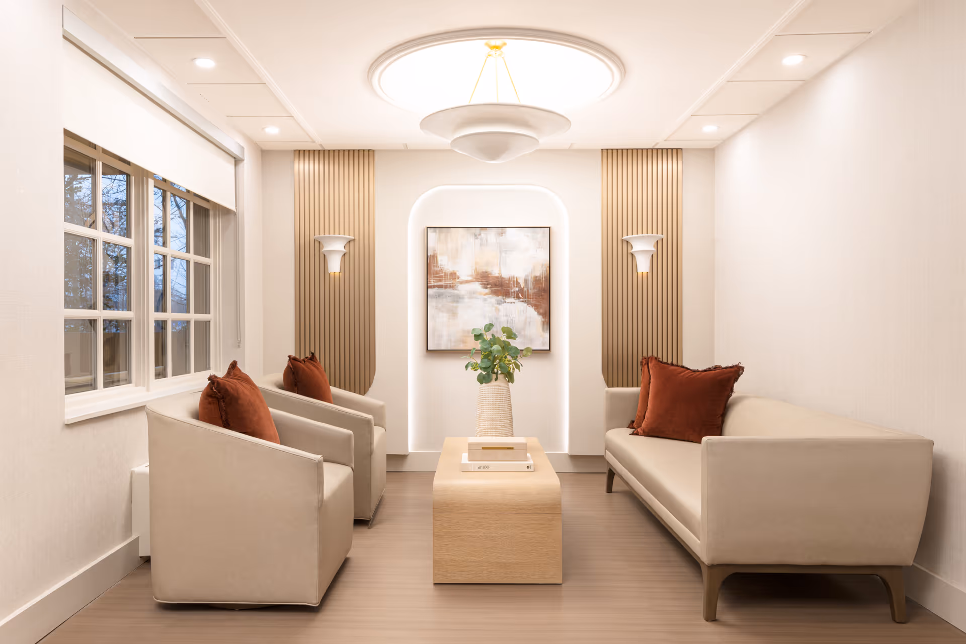 A modern and cozy living room with two beige armchairs and a beige sofa, each adorned with rust-colored throw pillows. A wooden coffee table with a vase of greenery and books sits in the center. The room features a large window on the left, soft lighting from ceiling fixtures, wall sconces, and a framed abstract painting on the back wall with vertical wooden panel accents.