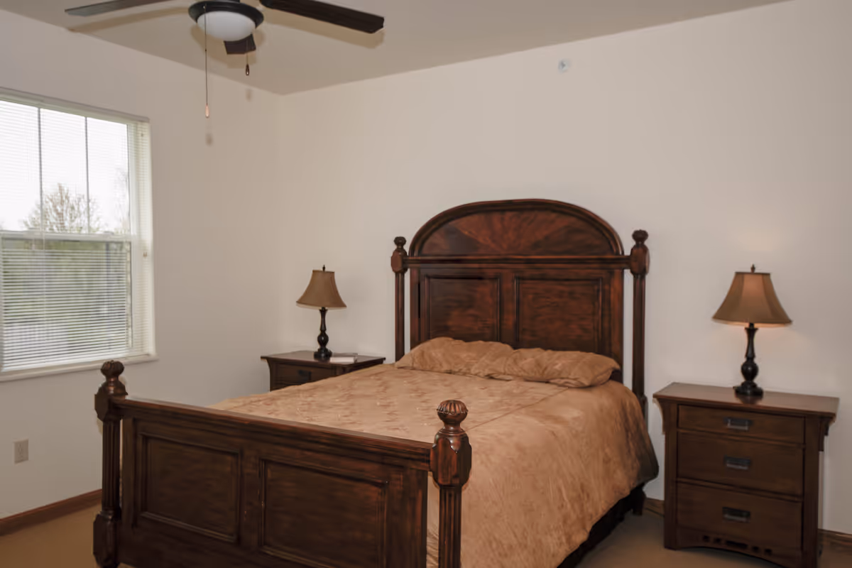 A bedroom with a large wooden bed featuring a high headboard and footboard, covered with a beige quilt. There are two matching wooden nightstands on either side of the bed, each with a lamp. A window with blinds is on the left wall, and a ceiling fan with a light fixture is visible above.